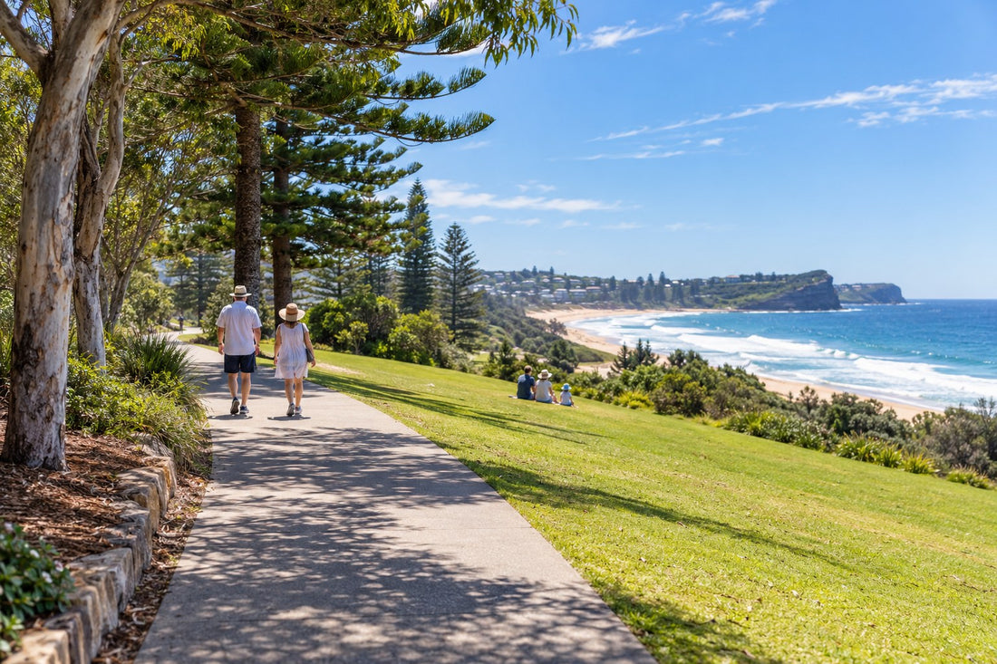 Sunlit Australian coastal park walkway at midday in summer with Norfolk pines, surf beach and walkers in sun hats