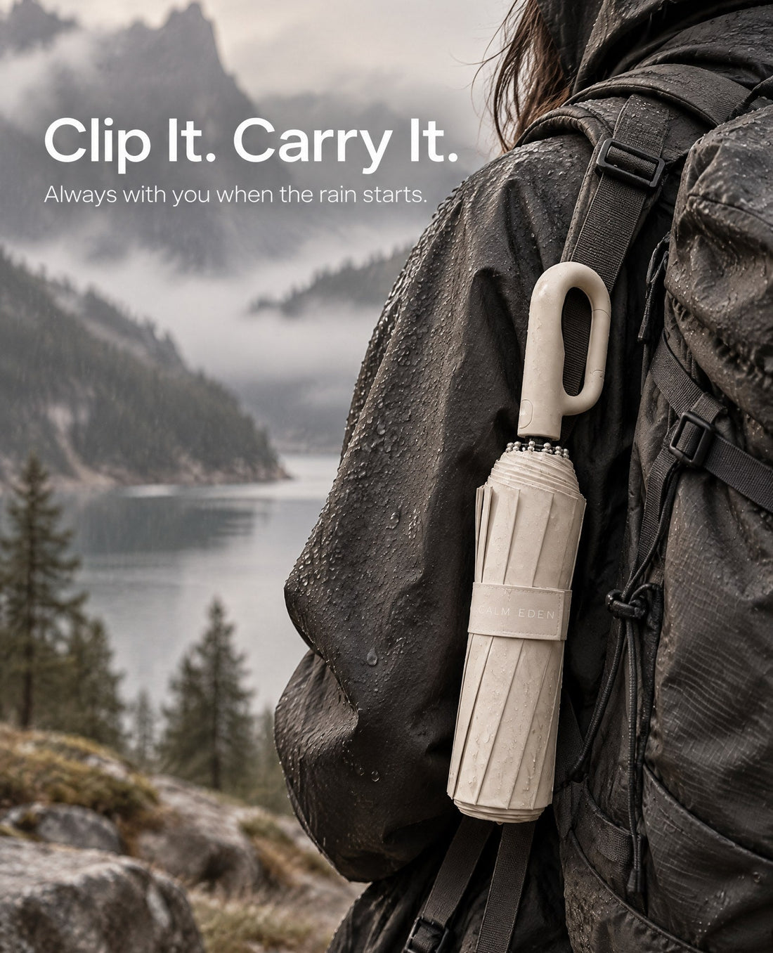 Haven compact umbrella clipped by its carabiner loop handle to the shoulder strap of a hiking backpack, with a misty lake and mountains behind
