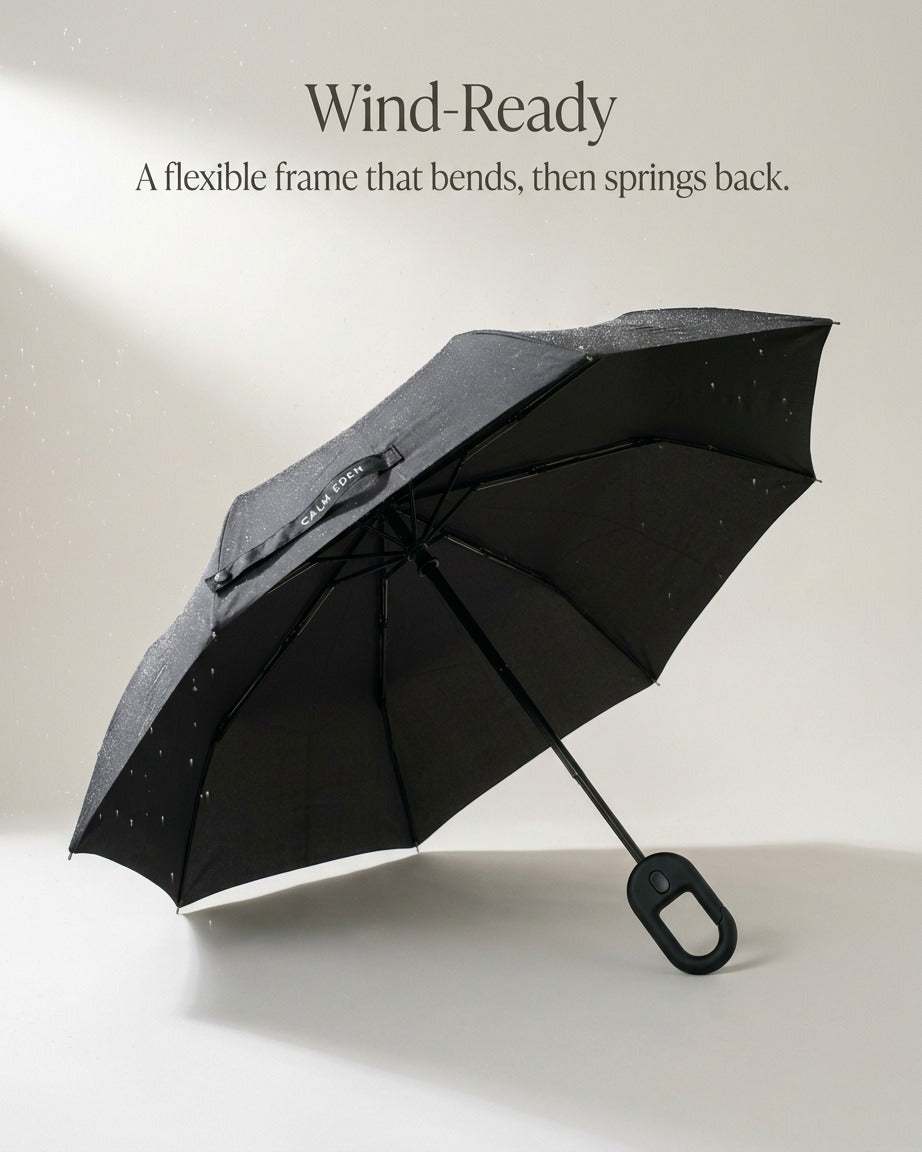 Open black Haven umbrella tilted on a pale studio floor, raindrops on the canopy and the ribs visible underneath, with the caption Wind-Ready: a flexible frame that bends, then springs back