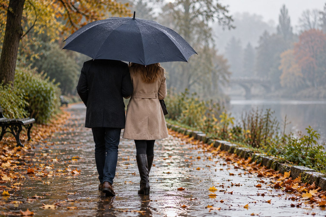Two people sharing one large navy golf umbrella on a wet park footpath covered in autumn leaves