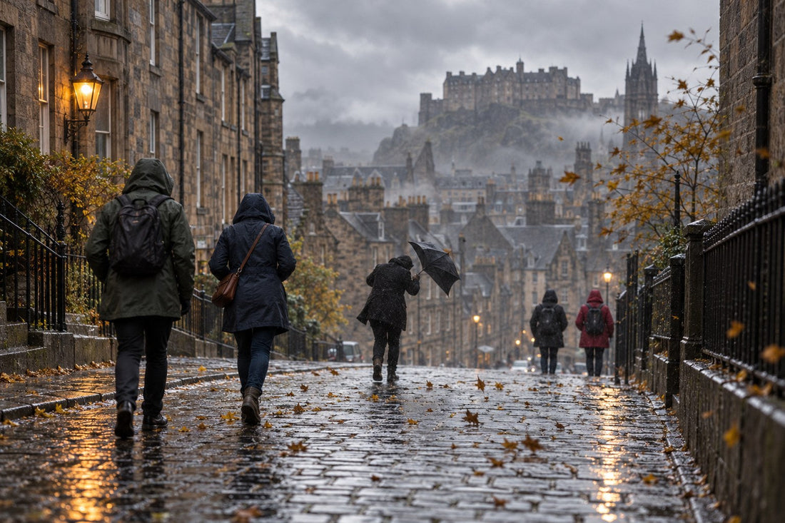 Wet cobbled old-town street in Scotland on a blustery grey afternoon with pedestrians in waterproof jackets and one dark umbrella bent by a gust