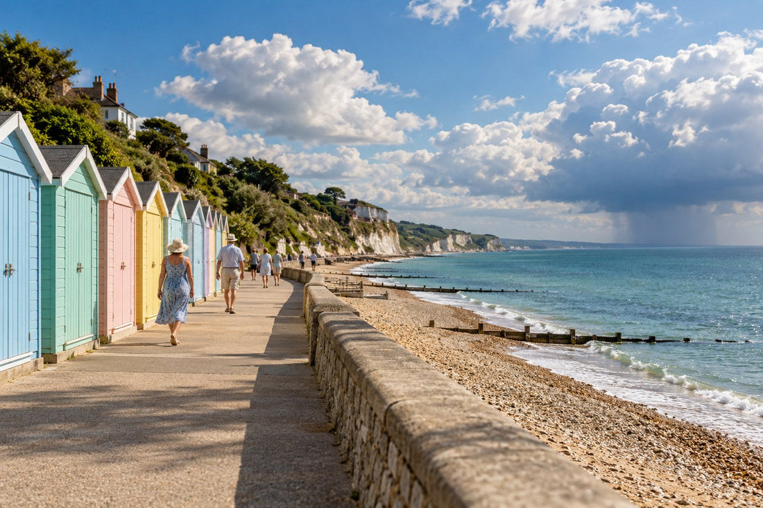 English seaside promenade with pastel beach huts on a bright summer afternoon, white cumulus clouds and a distant rain shower over the sea
