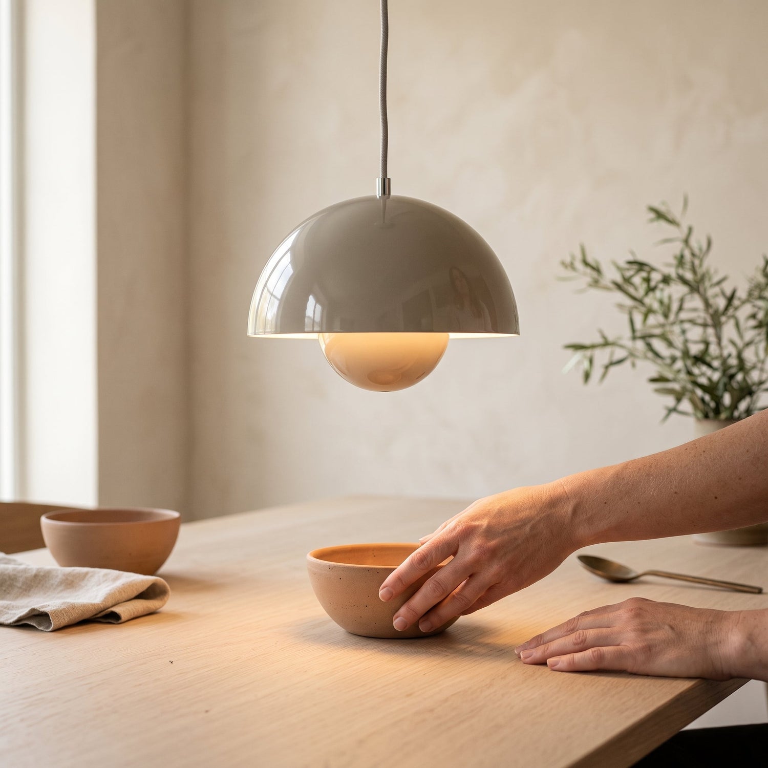 Hands setting a clay bowl on the table in the warm pool of light beneath the Bud pendant