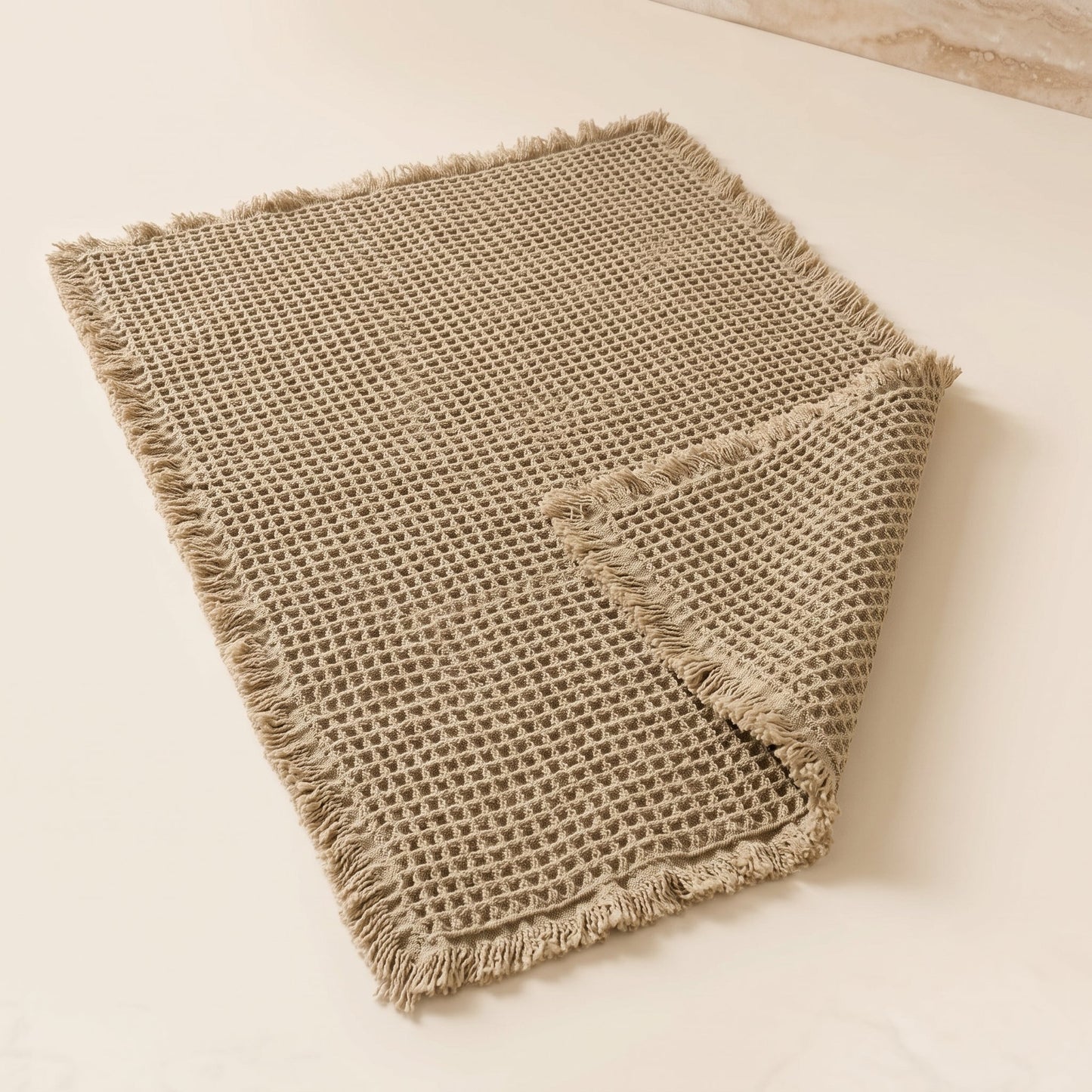 Waffle™ Bath Mat — greige waffle-weave mat with fringed ends on warm cream background