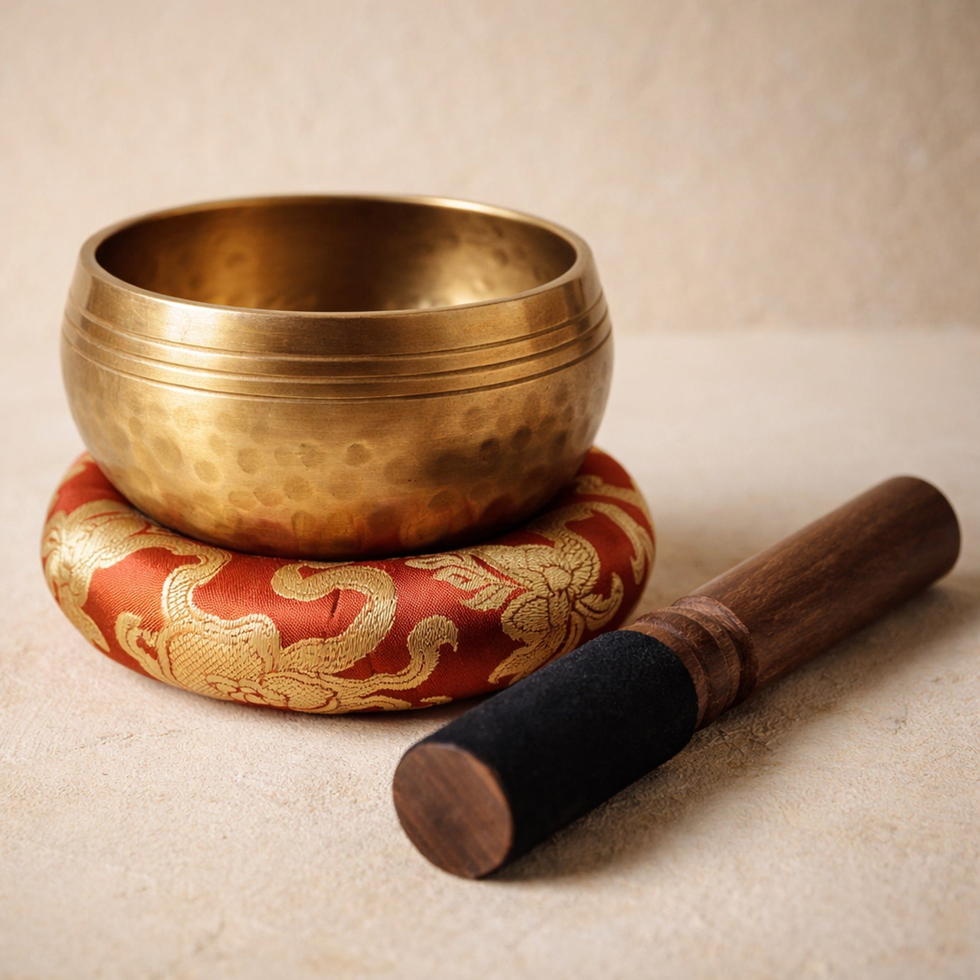 Chant™ Singing Bowl — dimpled copper-alloy bowl resting in its red brocade cushion with the suede-headed mallet, on warm cream plaster