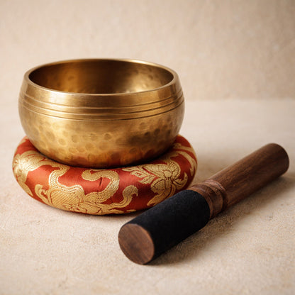 Chant™ Singing Bowl — dimpled copper-alloy bowl resting in its red brocade cushion with the suede-headed mallet, on warm cream plaster