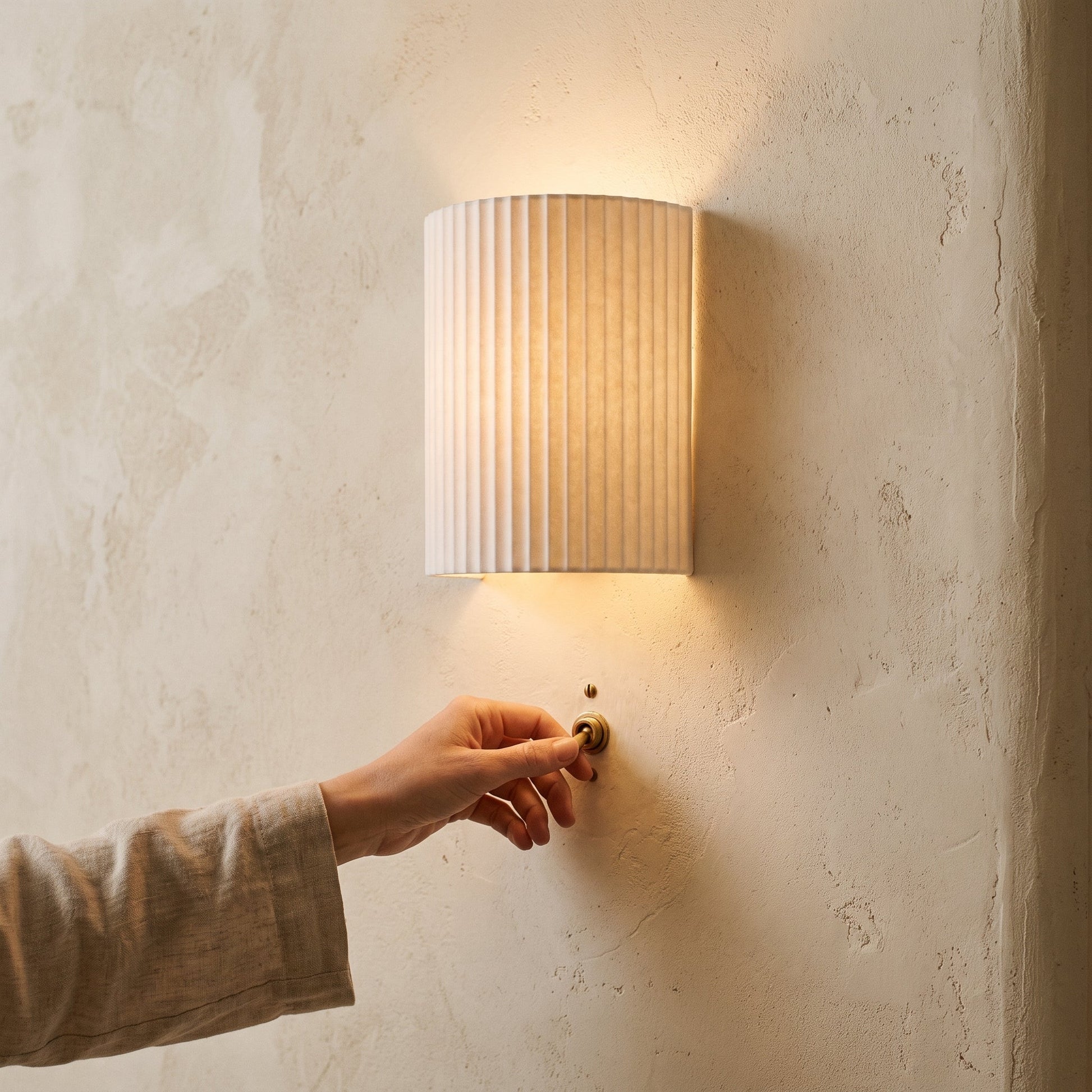 Hand reaching for the switch beneath a lit white Flute sconce at dusk